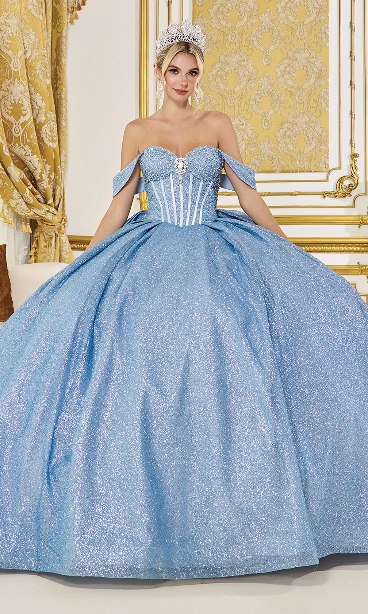 A woman in the Quinceanera Dress 1937 by Dancing Queen, featuring off-the-shoulder straps and glitter details, pairs it with a silver tiara as she sits in an ornate gold and white room.